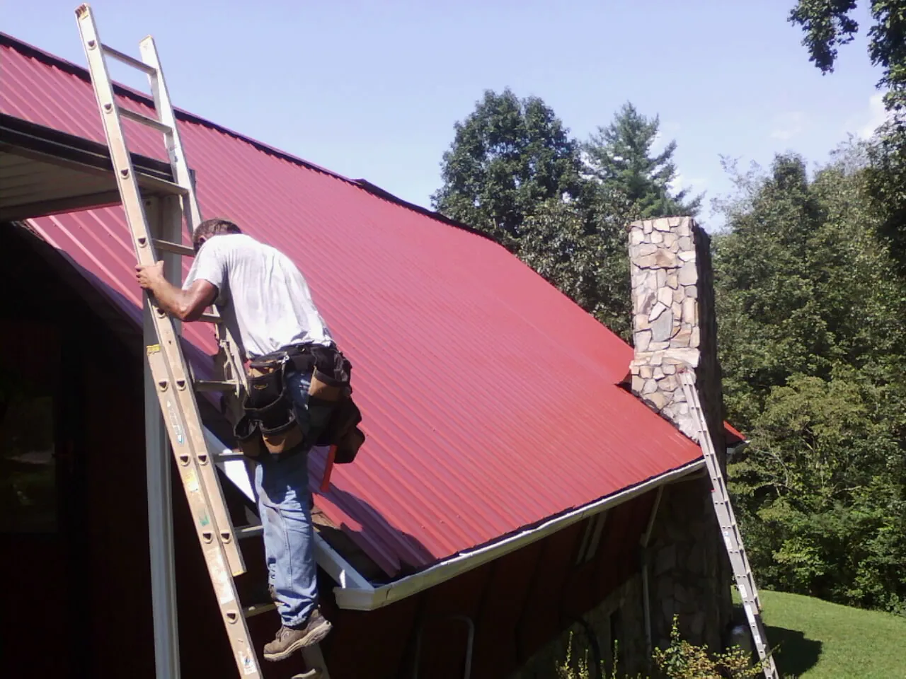 Roofer on a ladder beside a red metal roof and stone chimney — Soffit & Fascia Repair by Roofing Brothers in Ryderwood